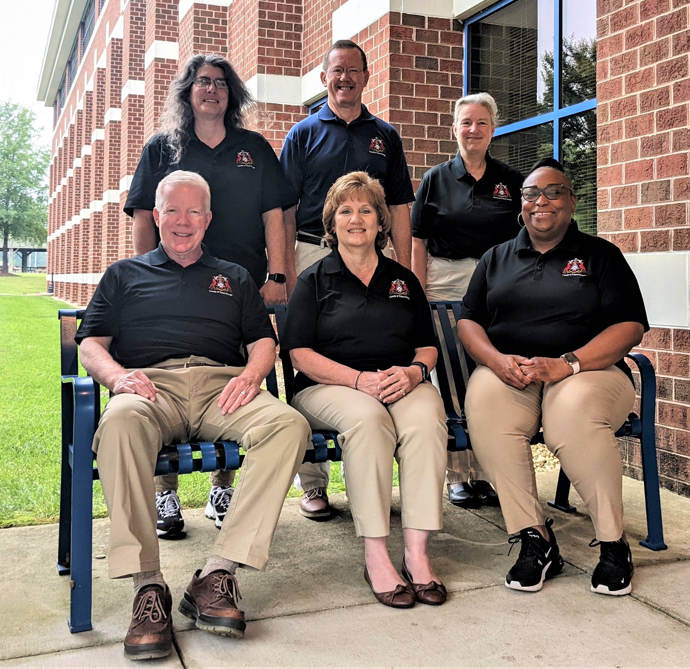 The photo depicts the Prince George County Real Estate Assessor's Team. There are six individuals in the photo. They are all smiling and wearing black polo shirts and tan pants. 